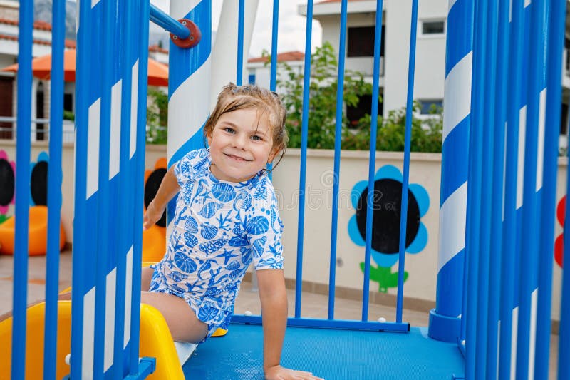 Little preschool girl sliding on a children slide in outdoor swimming pool in hotel resort. Child learning to swim in outdoor pool, splashing with water, laughing and having fun. Family vacations. Little preschool girl sliding on a children slide in outdoor swimming pool in hotel resort. Child learning to swim in outdoor pool, splashing with water, laughing and having fun. Family vacations