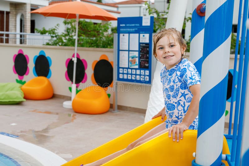 Little preschool girl sliding on a children slide in outdoor swimming pool in hotel resort. Child learning to swim in outdoor pool, splashing with water, laughing and having fun. Family vacations. Little preschool girl sliding on a children slide in outdoor swimming pool in hotel resort. Child learning to swim in outdoor pool, splashing with water, laughing and having fun. Family vacations