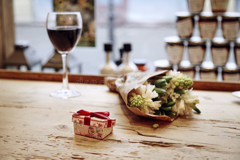 Liten gullig närvarande ask med pilbågen på trätabellen, blommor och exponeringsglas av vin bakom Romantiskt möte i kafé Dag c fö arkivfoton