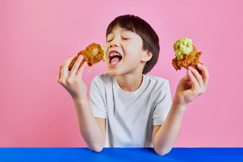 Little Korean boy, child in white t-shirt sitting at table and emotionally eating friend chicken with mustard sauce against pink background. Concept of food, childhood, emotions, meal, menu, pop art. Little Korean boy, child in white t-shirt sitting at table and emotionally eating friend chicken with mustard sauce against pink background. Concept of food, childhood, emotions, meal, menu, pop art