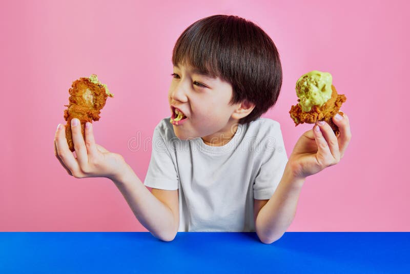 Little Korean boy, child in white t-shirt sitting at table and emotionally eating friend chicken with mustard sauce against pink background. Concept of food, childhood, emotions, meal, menu, pop art. Little Korean boy, child in white t-shirt sitting at table and emotionally eating friend chicken with mustard sauce against pink background. Concept of food, childhood, emotions, meal, menu, pop art