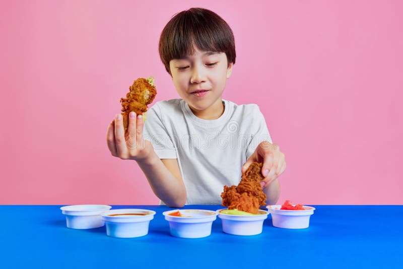 Little boy, Korean kid sitting at table and eating fried chicken, dipping in different sauces against pink background. Concept of food, childhood, emotions, meal, menu, pop art. Little boy, Korean kid sitting at table and eating fried chicken, dipping in different sauces against pink background. Concept of food, childhood, emotions, meal, menu, pop art