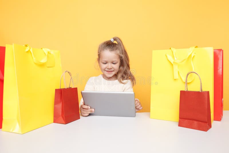 Liten leende flicka online med en bärbar dator och sitter på skrivbordet med shoppingväskor royaltyfri fotografi Liten leende flicka online med en bärbar dator och sitter på skrivbordet med shoppingväskor royaltyfri fotografi