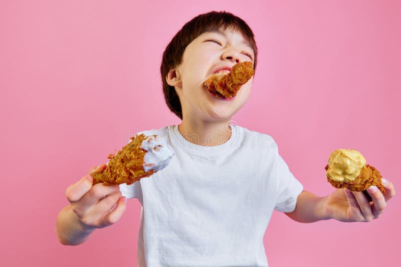 Little boy, child in white t-shirt emotionally eating friend chicken, making big bites against pink background. Fast food lover. Concept of food, childhood, emotions, meal, menu, pop art. Little boy, child in white t-shirt emotionally eating friend chicken, making big bites against pink background. Fast food lover. Concept of food, childhood, emotions, meal, menu, pop art