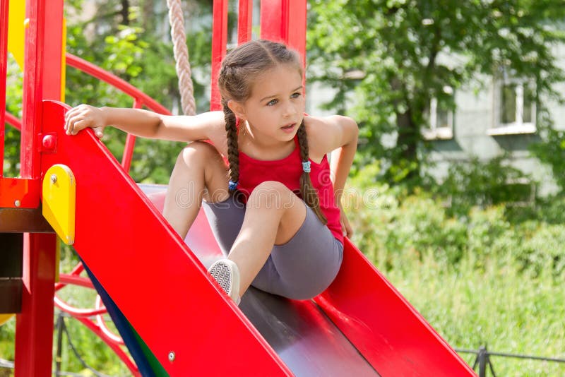 Liten sportive flicka som spelar på lekplatsen som sitter på glidbanan arkivbild Liten sportive flicka som spelar på lekplatsen som sitter på glidbanan arkivbild