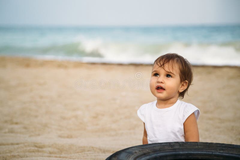 Litet behandla som ett barn, lilla flickan i den vita t-skjortan som nära sitter i sand på stranden till det gamla gummihjulet av royaltyfria foton Litet behandla som ett barn, lilla flickan i den vita t-skjortan som nära sitter i sand på stranden till det gamla gummihjulet av royaltyfria foton