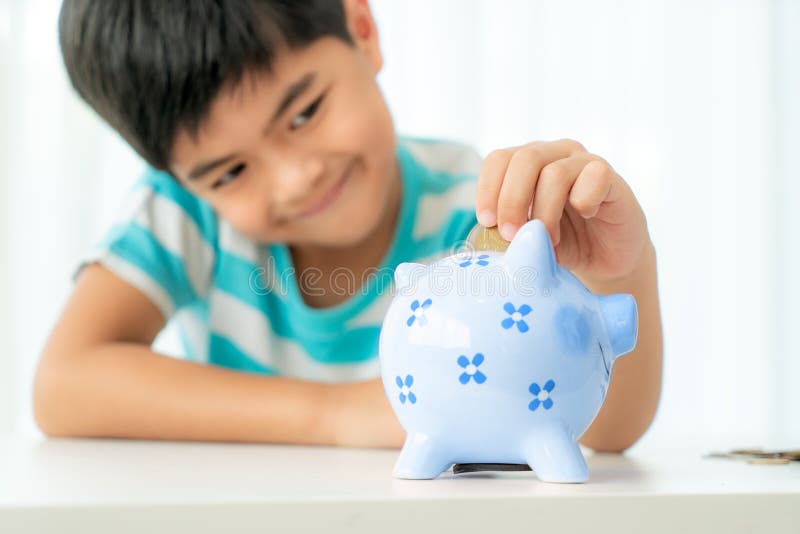 Little Asian boy insert a coin into blue piggy bank in white table at living room at home for the kid saving, investment money for