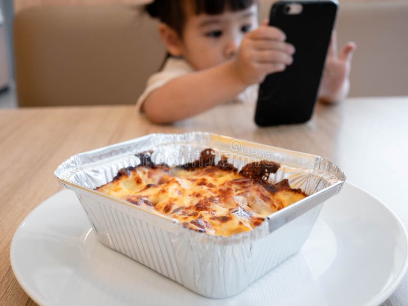 Little Asian child girl eating lunch while watching cartoon on the smartphone in a restaurant with her family. Child addicted to. Mobile phones stock photos