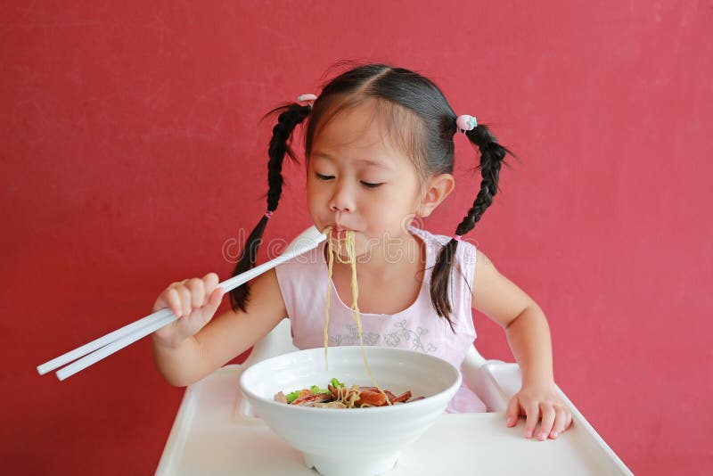 Little asian girl eating Egg noodle with roasted duck and pork on high chair against red wall background