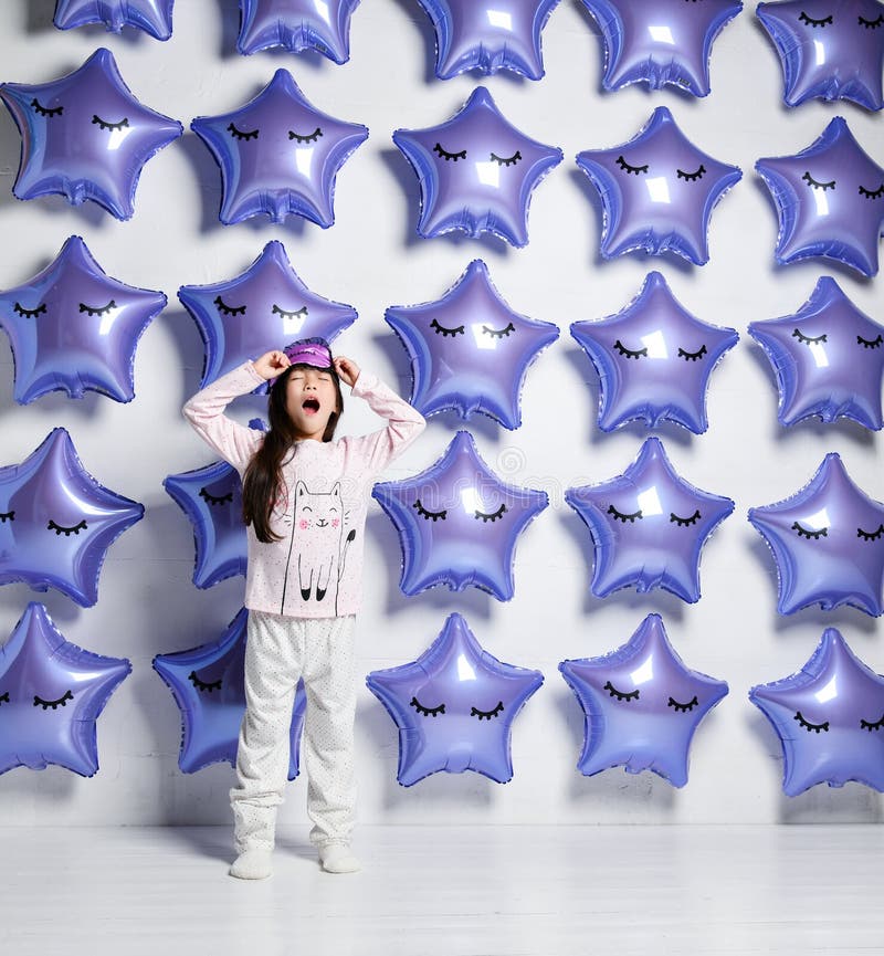 Little asian girl kid in pyjamas yawns and pulls on the eye mask for sleep on background of big balloons stars royalty free stock photography Little asian girl kid in pyjamas yawns and pulls on the eye mask for sleep on background of big balloons stars royalty free stock photography