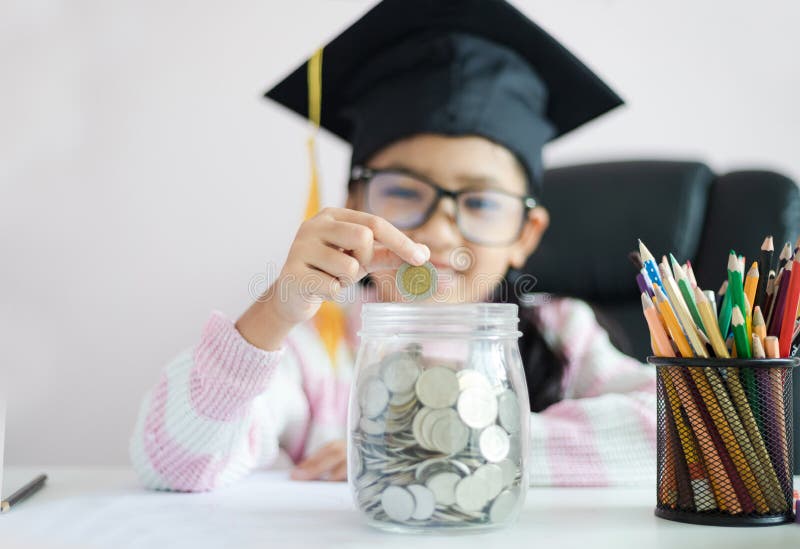 Little Asian girl wearing graduate hat putting the coin into clear glass jar piggy bank and smile with happiness for money saving