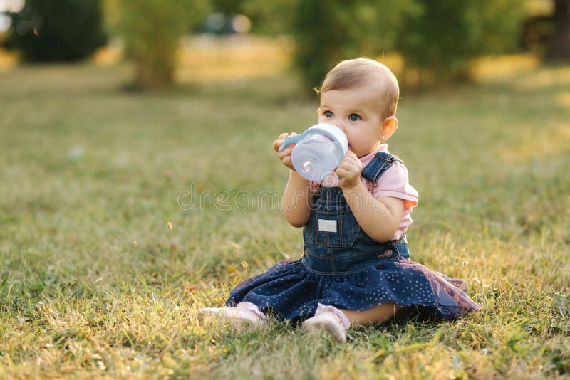 Little baby drink water from baby bottle outside in the park. Baby gir sit on the grass