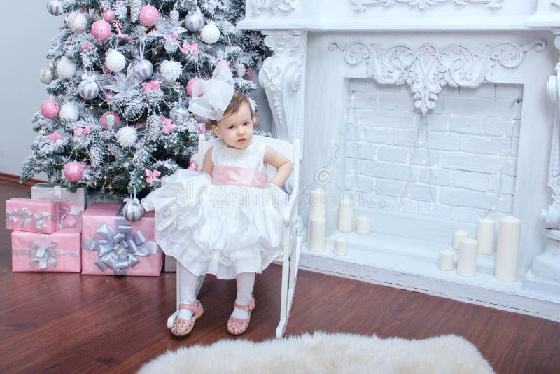 A little beautiful girl two years old dressed in a magnificent white dress with a pink bow is sitting in a chair near an elegant stock photo A little beautiful girl two years old dressed in a magnificent white dress with a pink bow is sitting in a chair near an elegant stock photo