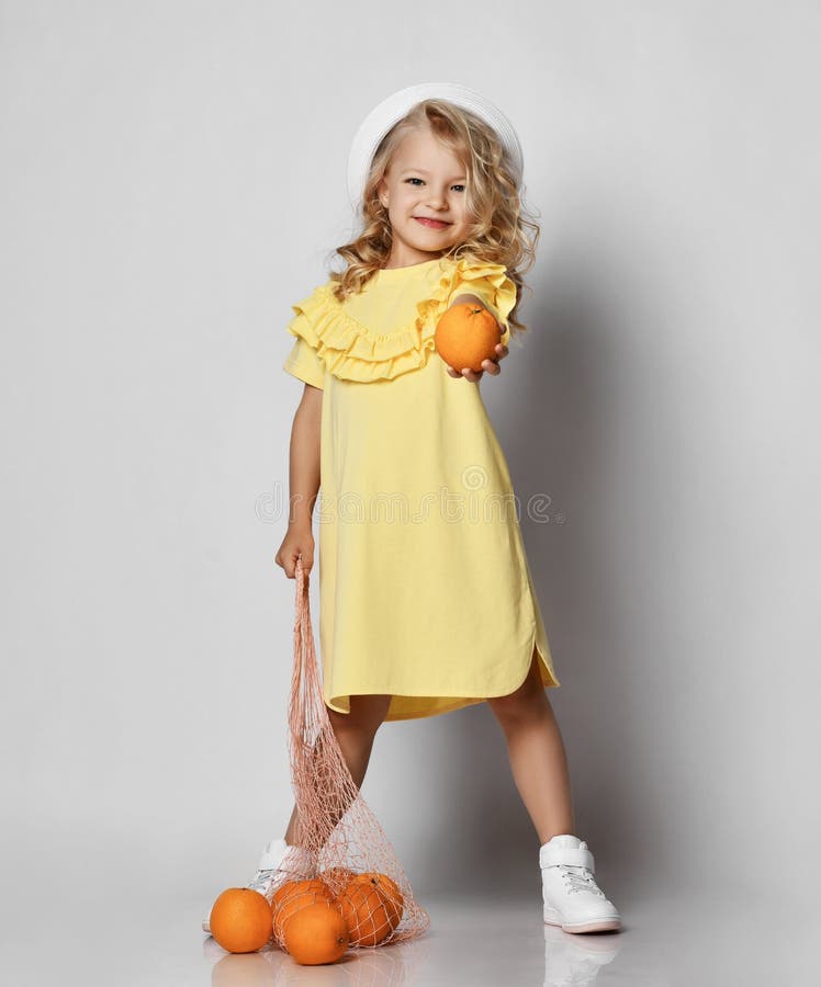 Little blonde curly princess girl in yellow dress, sneakers and hat standing and holding bag with oranges
