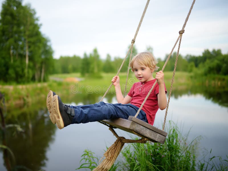 Little boy having fun on a swing hanging on big tree near pond or river in the forest in summer day