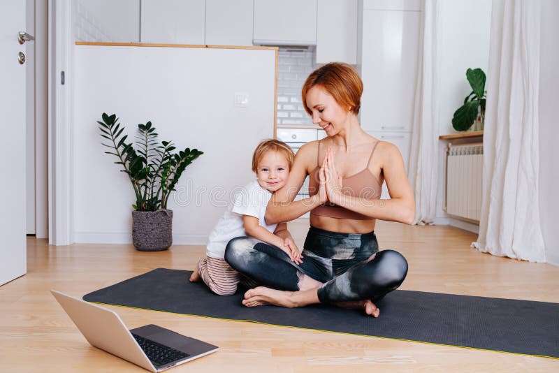 Happy little boy hugs his mother, while doing yoga on a yoga mat in front of a laptop at home. She`s looking at her son. She wears tight sportive clothes. Happy little boy hugs his mother, while doing yoga on a yoga mat in front of a laptop at home. She`s looking at her son. She wears tight sportive clothes