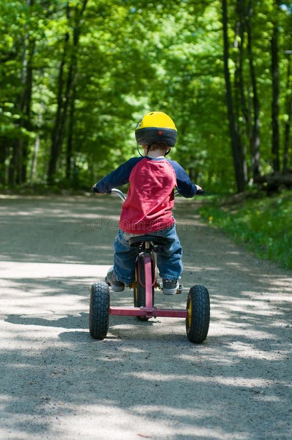 A young boy riding a tricycle away from the camera down a country lane. A young boy riding a tricycle away from the camera down a country lane