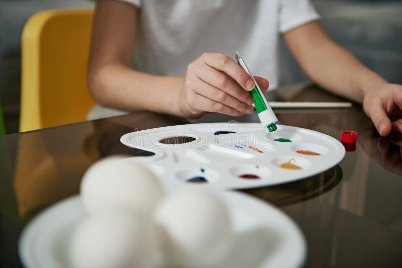 Little boy`s hand squeezing green paint from a tube onto a palette, blurry eggs on a saucer in the foreground. Closeup