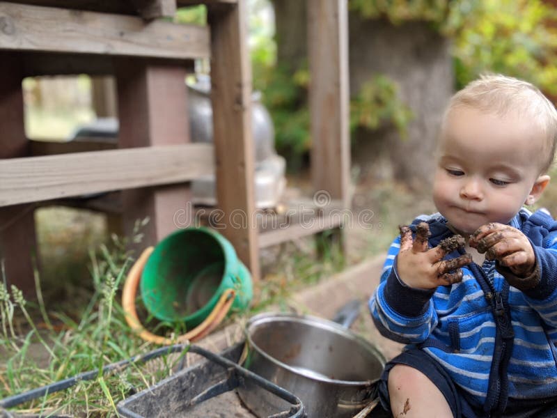 Little boy sitting on the ground looking at and picking his muddy hand