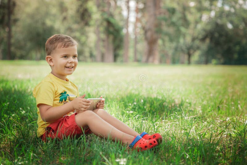 A little boy is sitting with the phone on the grass.