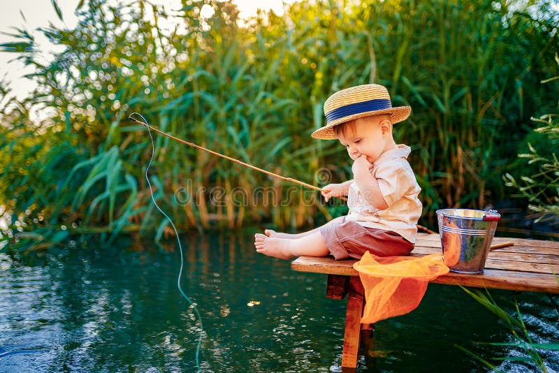 Little boy in straw hat sitting on the edge of a wooden dock and fishing in lake at sunset