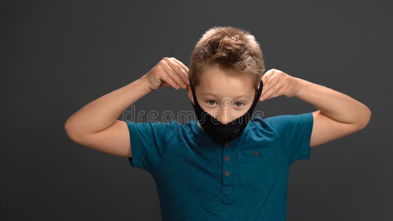 Little boy takes off his black mask with his hand on gray background. Caucasian child breathes with relief and smiles looking at camera. Concept of ending pandemic.