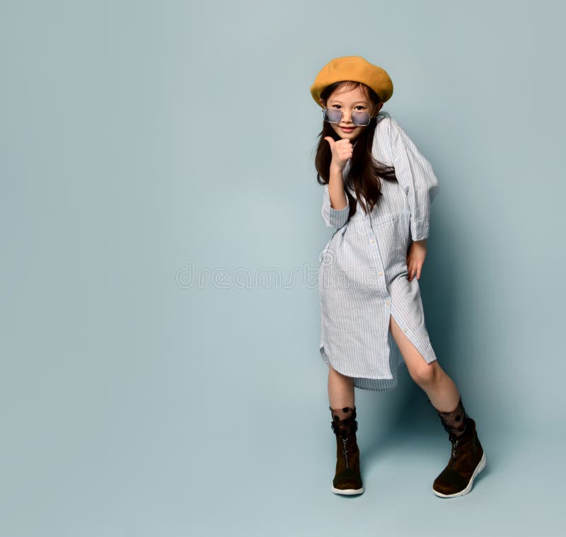 Little asian kid in sunglasses, oversized shirt dress, brown beret, boots. She kissing you, posing on blue background. Full length