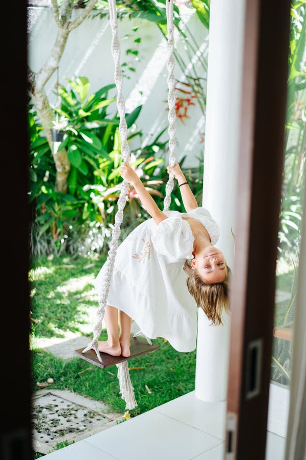 Little brunette girl standing on swings in a courtyard, bowing to look back