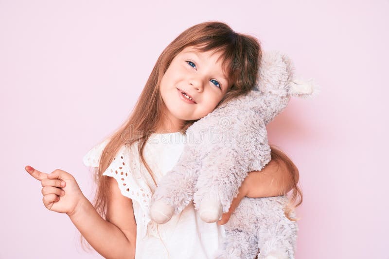 Little caucasian kid girl with long hair holding stuffed animal smiling happy pointing with hand and finger to the side