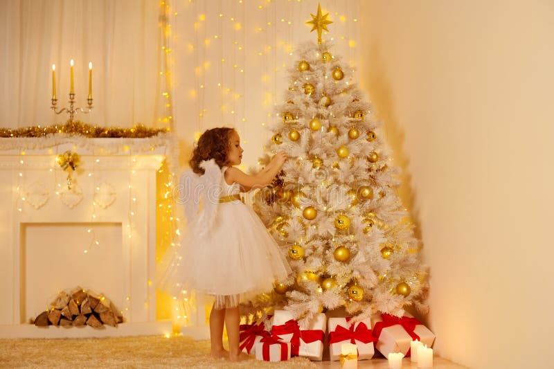 Little Child Girl Decorating Christmas Tree with Golden Ball in Cozy White Interior with Fireplace. Xmas Room Lights