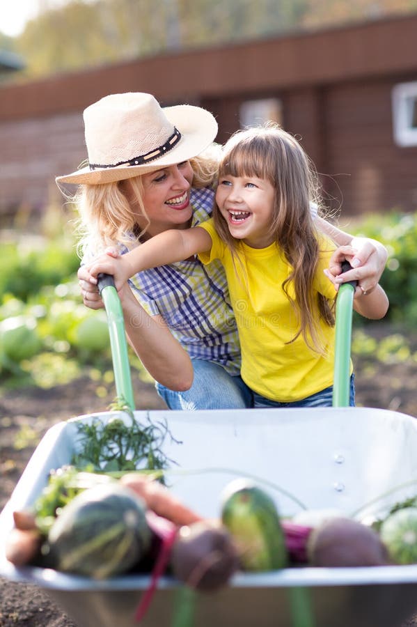 Woman Pushing Her Child Daughter In A Wheelbarrow Filled Vegetables In