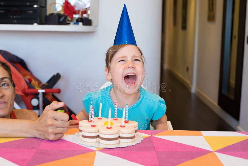 Three years old blonde child with blue shirt and cone hat, and birthday cake with candles on colorful tablecloth, screaming angry, next to women with lighter in hand. Three years old blonde child with blue shirt and cone hat, and birthday cake with candles on colorful tablecloth, screaming angry, next to women with lighter in hand
