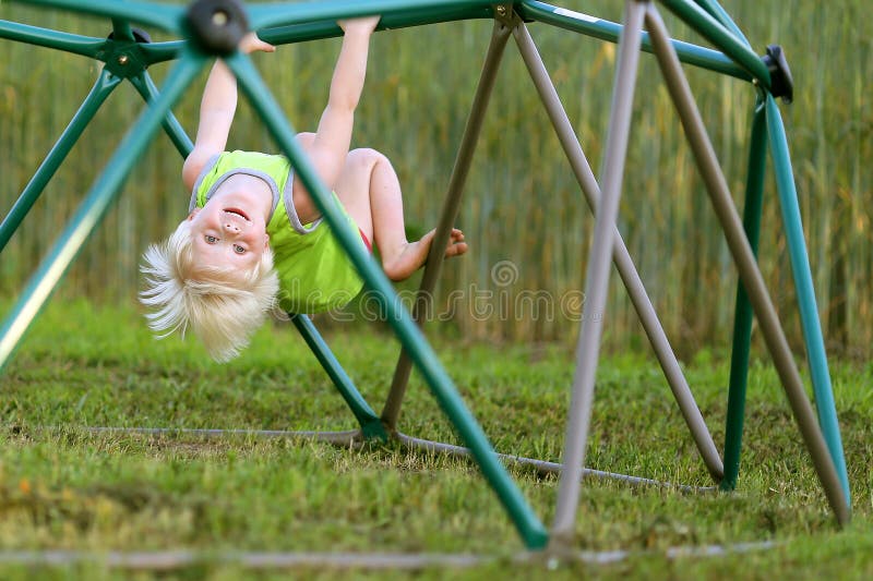 A little boy child with light blond hair is smiling as he hangs upside down while cli...