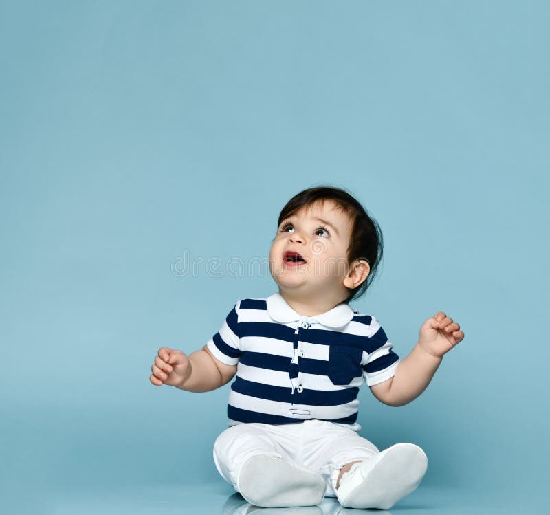 Little child in striped t-shirt, white pants and booties. He is looking up, sitting on floor against blue background. Close up. Little child in striped t-shirt stock photo Little child in striped t-shirt, white pants and booties. He is looking up, sitting on floor against blue background. Close up. Little child in striped t-shirt stock photo