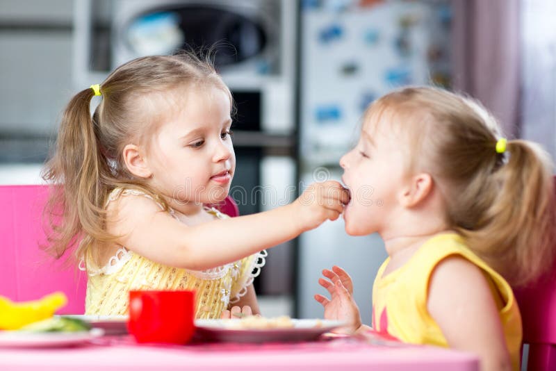 Little children toddlers eating meal together, one girl feeding sister in sunny kitchen at home