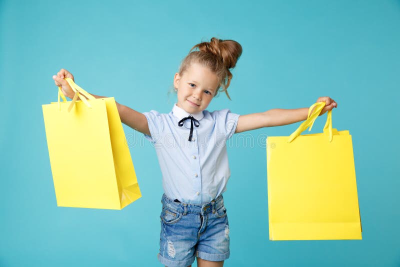 Little cute and funny girl holding big bright colored yellow paper bags in the blue studio. royalty free stock image Little cute and funny girl holding big bright colored yellow paper bags in the blue studio. royalty free stock image