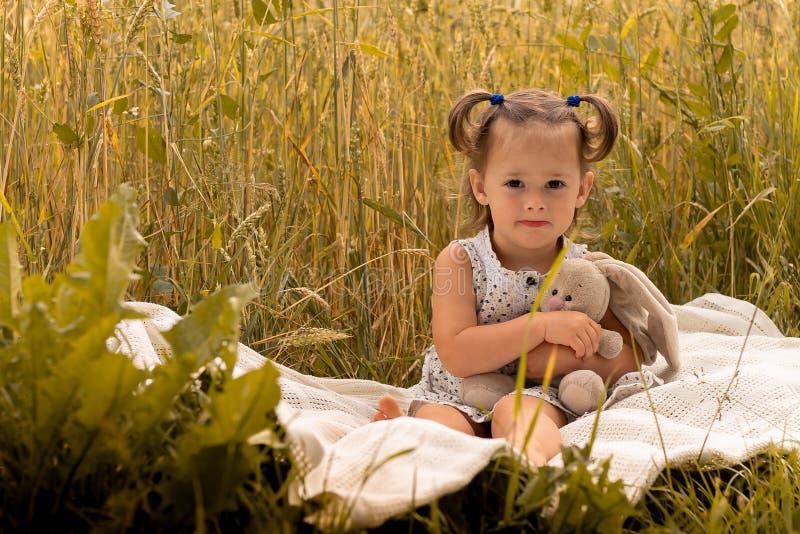 Little cute girl 1-3 in a light dress hugs a teddy hare sitting on a plaid in a field of spikelets of rye