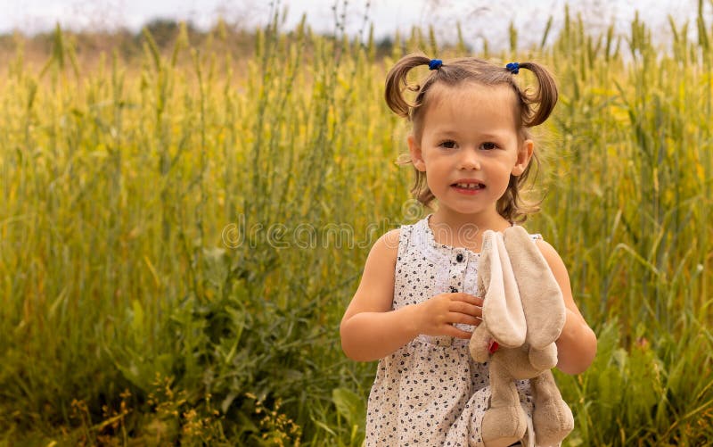 Little cute girl with a plush hare in a light dress 1-3 in the field of spikelets of rye