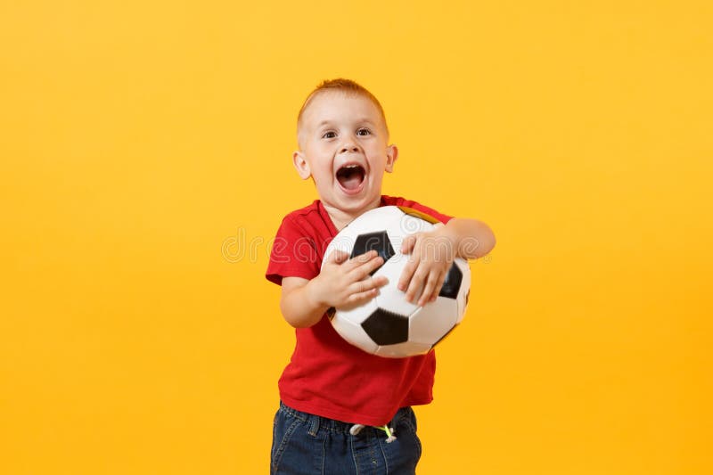 Little cute kid baby boy 3-4 years old, football fan in red t-shirt holding in hand soccer ball isolated on yellow