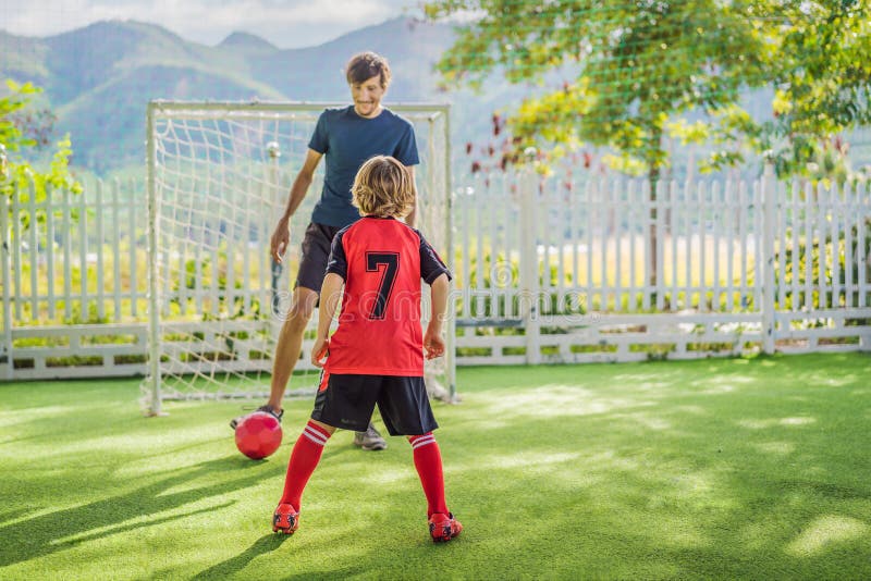 Little cute kid boy in red football uniform and his trainer or father playing soccer, football on field, outdoors