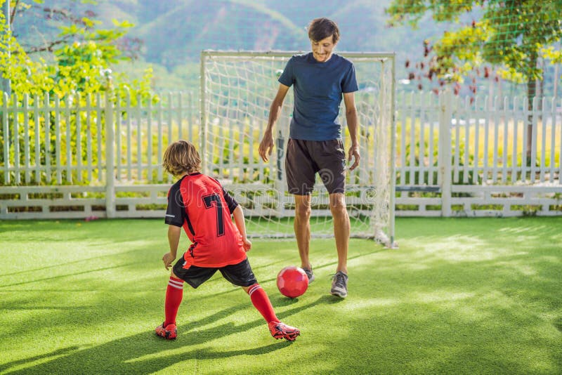 Little cute kid boy in red football uniform and his trainer or father playing soccer, football on field, outdoors