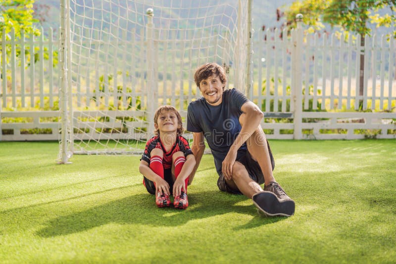 Little cute kid boy in red football uniform and his trainer or father playing soccer, football on field, outdoors