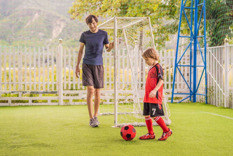 Little cute kid boy in red football uniform and his trainer or father playing soccer, football on field, outdoors