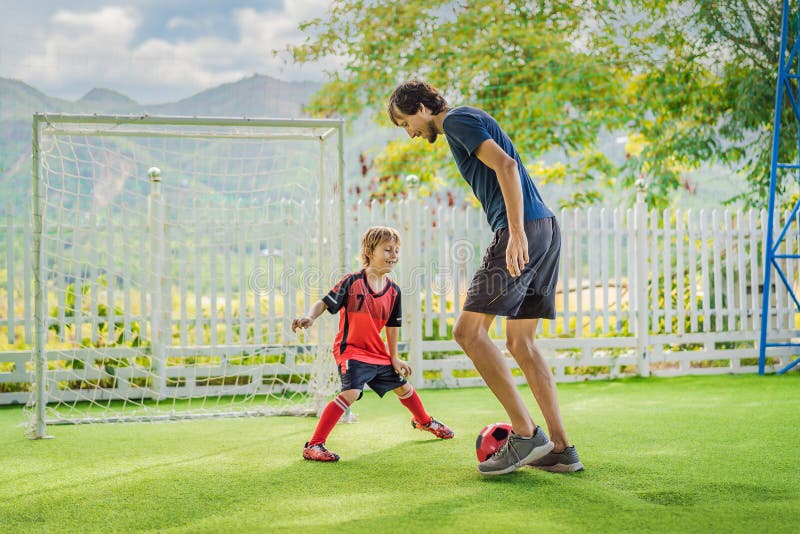 Little cute kid boy in red football uniform and his trainer or father playing soccer, football on field, outdoors