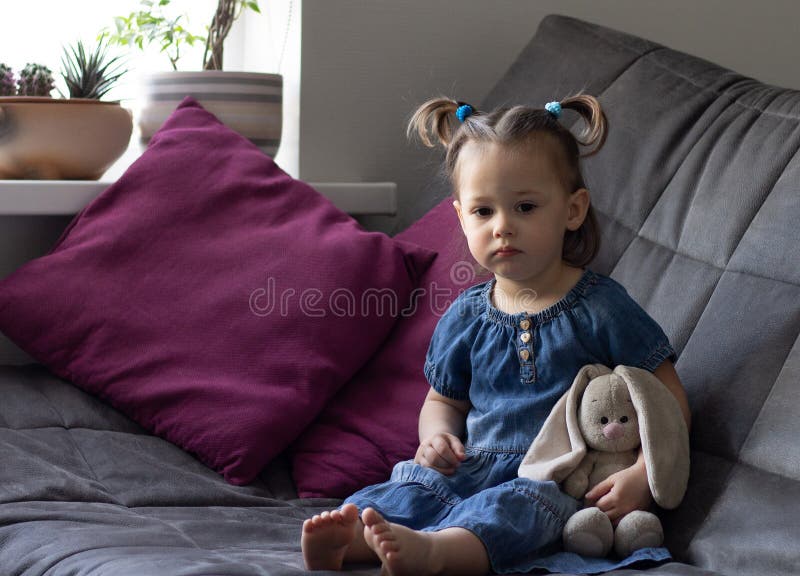 Little cute sad girl 1-3 is sitting on the sofa with a toy plush rabbit