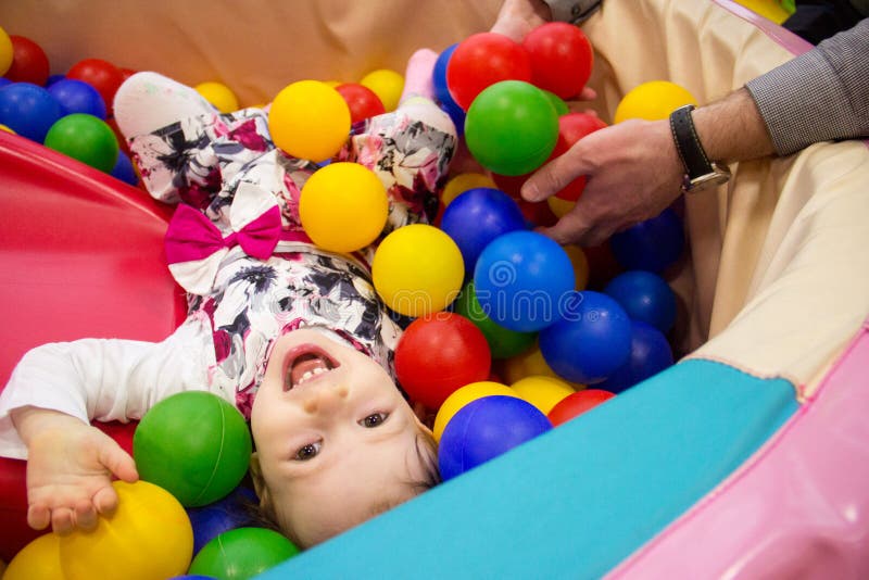 Little cute smile girl plays in balls for a dry pool. Play room. Happiness. father`s hand in the background stock photos Little cute smile girl plays in balls for a dry pool. Play room. Happiness. father`s hand in the background stock photos
