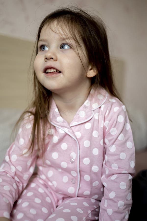 Little Dark-haired Girl in Pink Pajamas is Sitting on the Bed