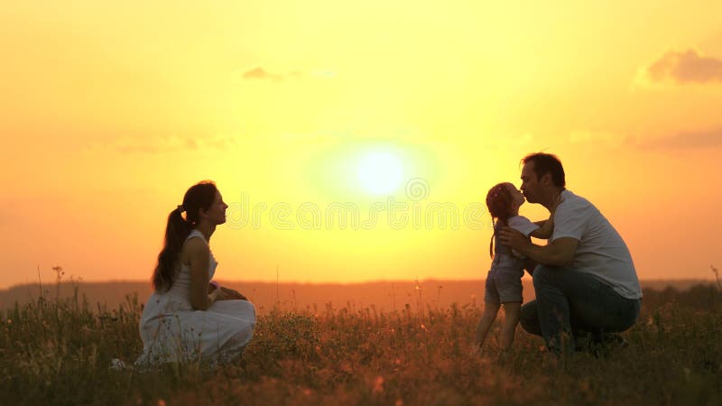 Little daughter goes from mom to dad, hugs and kisses her parents in rays of warm sun. Happy family walks in park at sunset. Mom, dad and baby. healthy family plays in field. Happy family concept. Little daughter goes from mom to dad, hugs and kisses her parents in rays of warm sun. Happy family walks in park at sunset. Mom, dad and baby. healthy family plays in field. Happy family concept