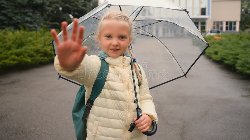 Little European girl wave hand at camera walk skipping umbrella city outside park schoolgirl jumping learner backpack child cheerful enjoy funny rainy weather daytime back to school greeting childish. High quality 4k footage. Little European girl wave hand at camera walk skipping umbrella city outside park schoolgirl jumping learner backpack child cheerful enjoy funny rainy weather daytime back to school greeting childish. High quality 4k footage