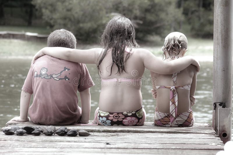 3 kleine Freunde, zwei Mädchen und ein junge, sitzen am Ende des dock, Blick auf das Wasser des Sees.